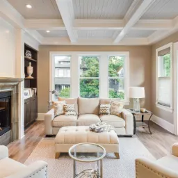 Jasny salon z beżową sofą, pufą i drewnianą podłogą, widok na okno z zielenią, sufit z białymi belkami i wbudowanym oświetleniem, oraz kominek z szarą obudową.