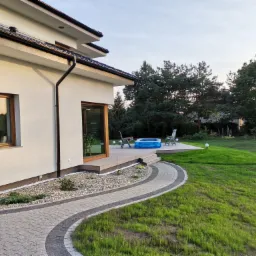 Nowoczesny dom z kremową elewacją, drewnianą stolarką okienną i tarasem z basenem dla dzieci, widok z boku na kamienny chodnik i trawnik.