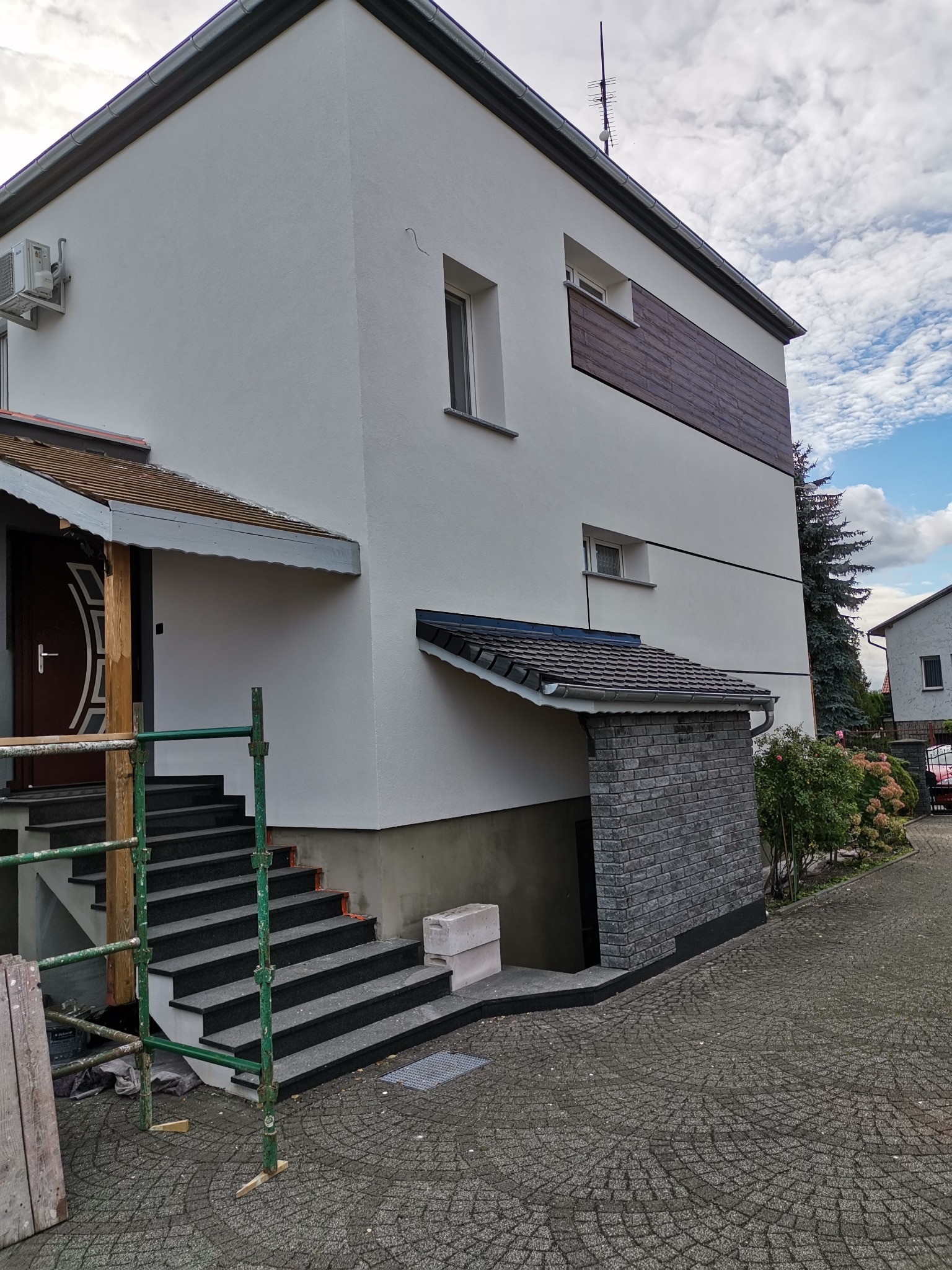 Nowoczesny dom murowany z białą elewacją, drewnianymi akcentami, schodami wejściowymi z czarnego granitu i ocieplonym wejściem do piwnicy wykończonym kamieniem elewacyjnym, częściowo zasłonięty...