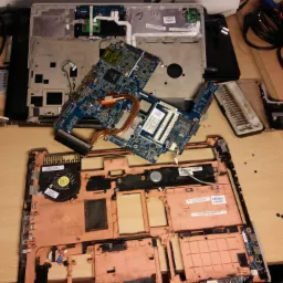 Rozłożony laptop na stole roboczym, widoczne płyty główne, wentylator, obudowa, śrubokręty i drobne śrubki.