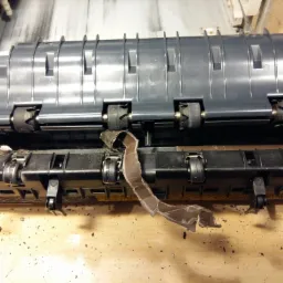 Wnętrze uszkodzonej drukarki z zablokowanym papierem i widocznymi elementami mechanizmu podawania papieru.