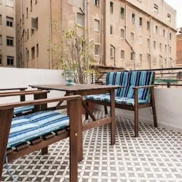 Umeblowany balkon z geometryczną posadzką, drewnianymi meblami i widokiem na budynki mieszkalne.