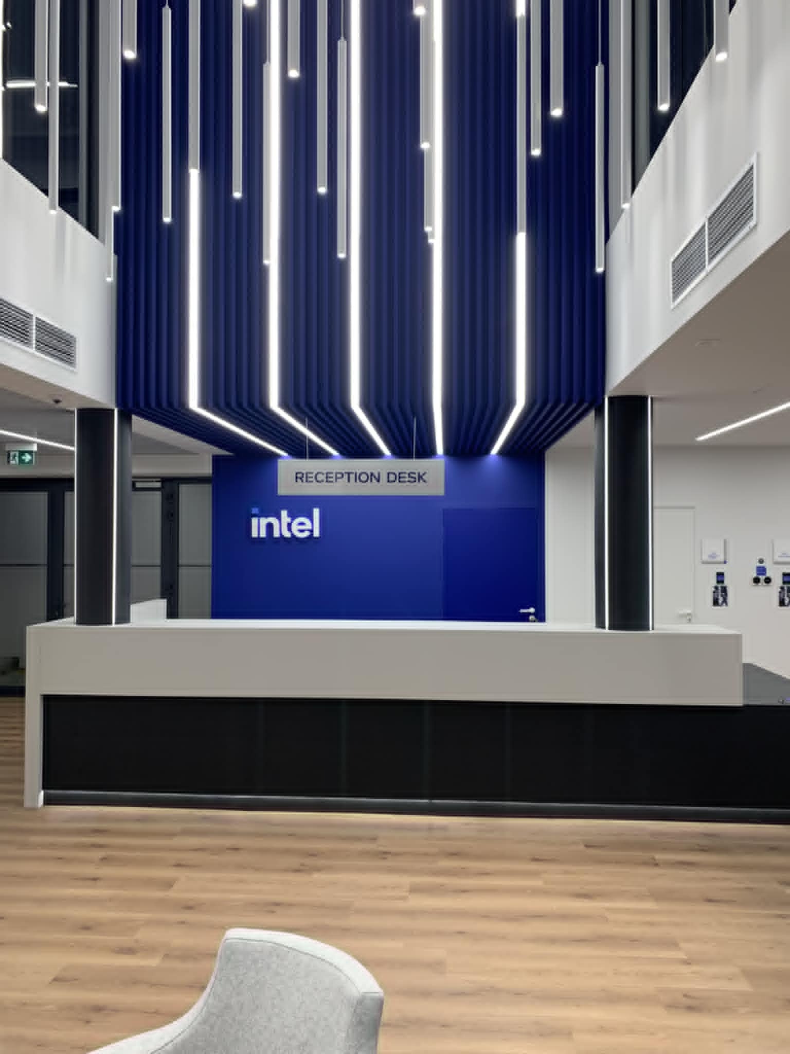Nowoczesne biuro z recepcją Intel, dominująca biel i błękit, podwieszany sufit z pionowymi lampami. Parkiet na podłodze, widoczne krzesło dla gości.