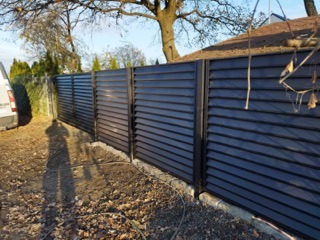 Nowoczesne ogrodzenie panelowe z poziomych, ciemnych listew, zamontowane na niskiej, kamiennej podmurówce. Widoczny cień osoby fotografującej oraz fragment białego samochodu dostawczego w tle.