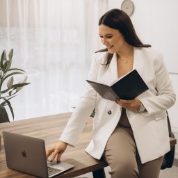 Biuro Nieruchomości Beinvest Poznań - Uśmiechnięta agentka nieruchomości w białym żakiecie, siedzi na biurku z laptopem i notatnikiem w jasnym biurze. Widok z boku, neutralne kolory.