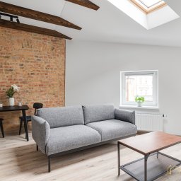 Biuro Nieruchomości Beinvest Poznań - Salon z szarą sofą, ceglaną ścianą i drewnianymi belkami. Widoczne okno dachowe, stół z krzesłami i stolik kawowy. Jasne wnętrze, minimalistyczny design.