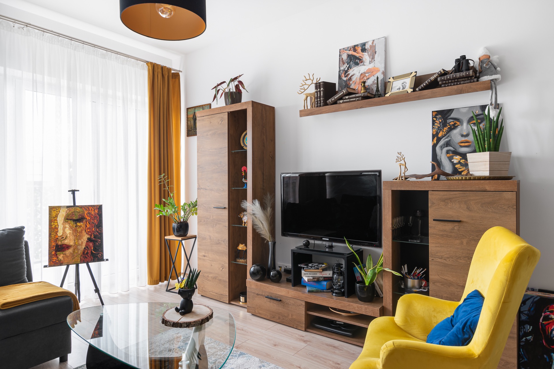 Jasny salon z drewnianymi meblami, żółtym fotelem i szklanym stolikiem kawowym, dodatkowo ozdobiony obrazami i roślinami doniczkowymi.