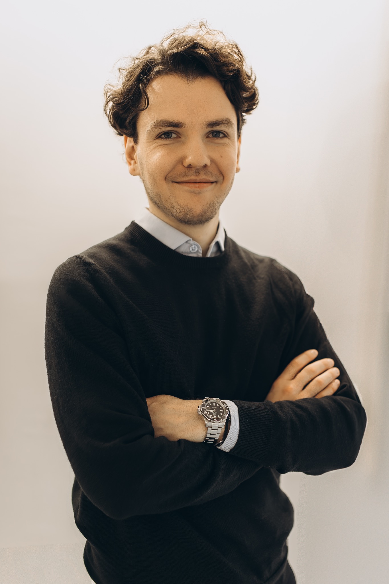Portret uśmiechniętego mężczyzny w czarnym swetrze i koszuli, z zegarkiem na nadgarstku, na jasnym tle. Skrzyżowane ręce.
