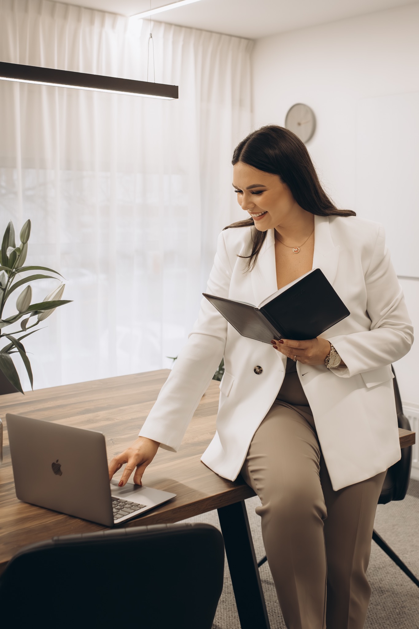 Uśmiechnięta agentka nieruchomości w białym żakiecie, siedzi na biurku z laptopem i notatnikiem w jasnym biurze. Widok z boku, neutralne kolory.