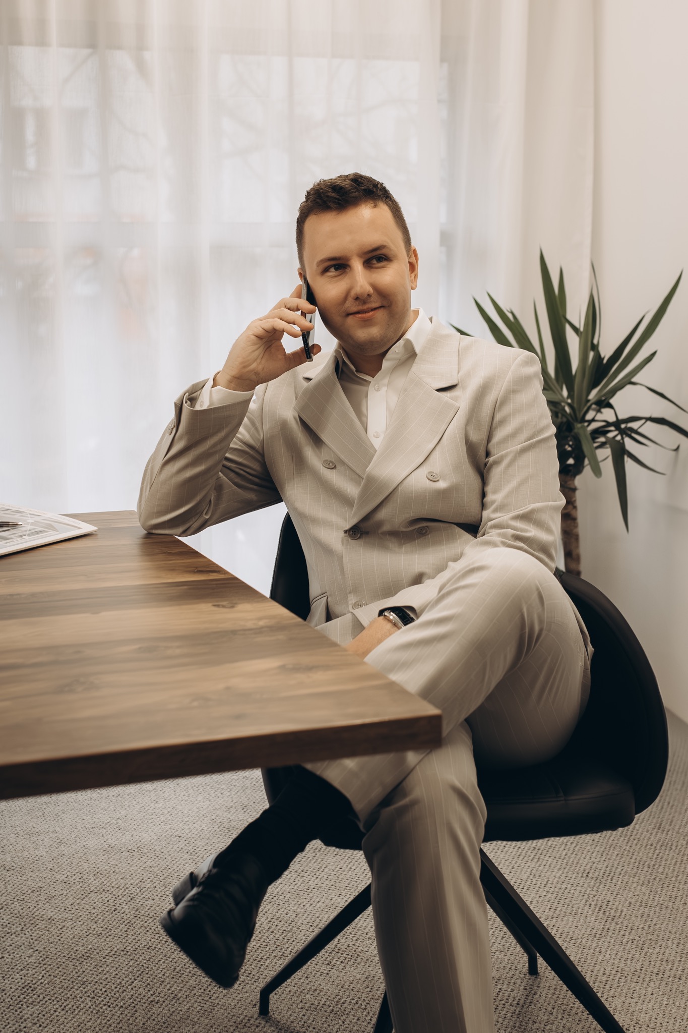 Mężczyzna w beżowym garniturze w paski siedzi przy biurku, rozmawiając przez telefon. W tle okno i roślina doniczkowa.