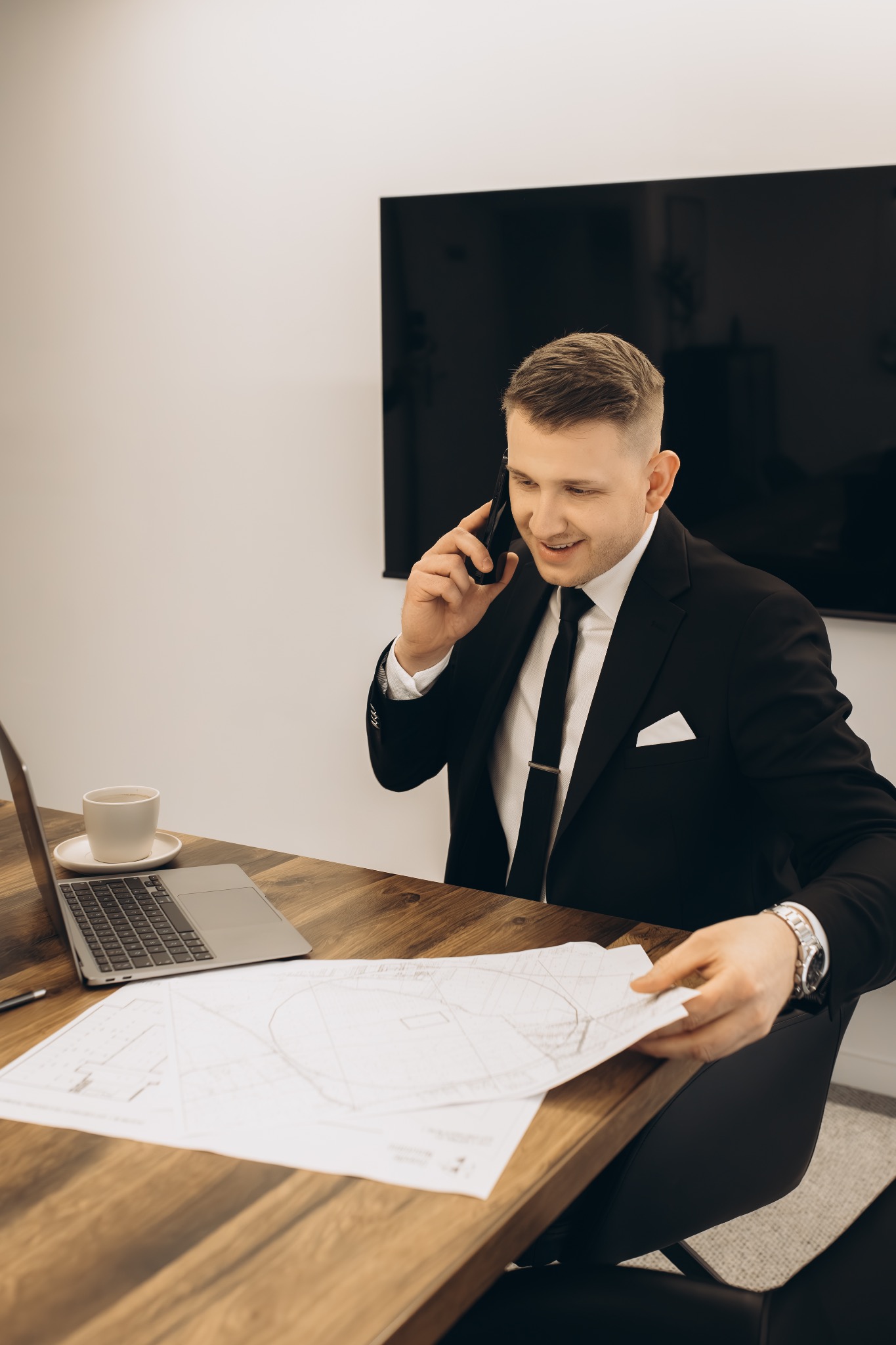 Zarządca nieruchomości w garniturze, rozmawia przez telefon, analizując plan nieruchomości na biurku z laptopem i kawą w nowoczesnym biurze.