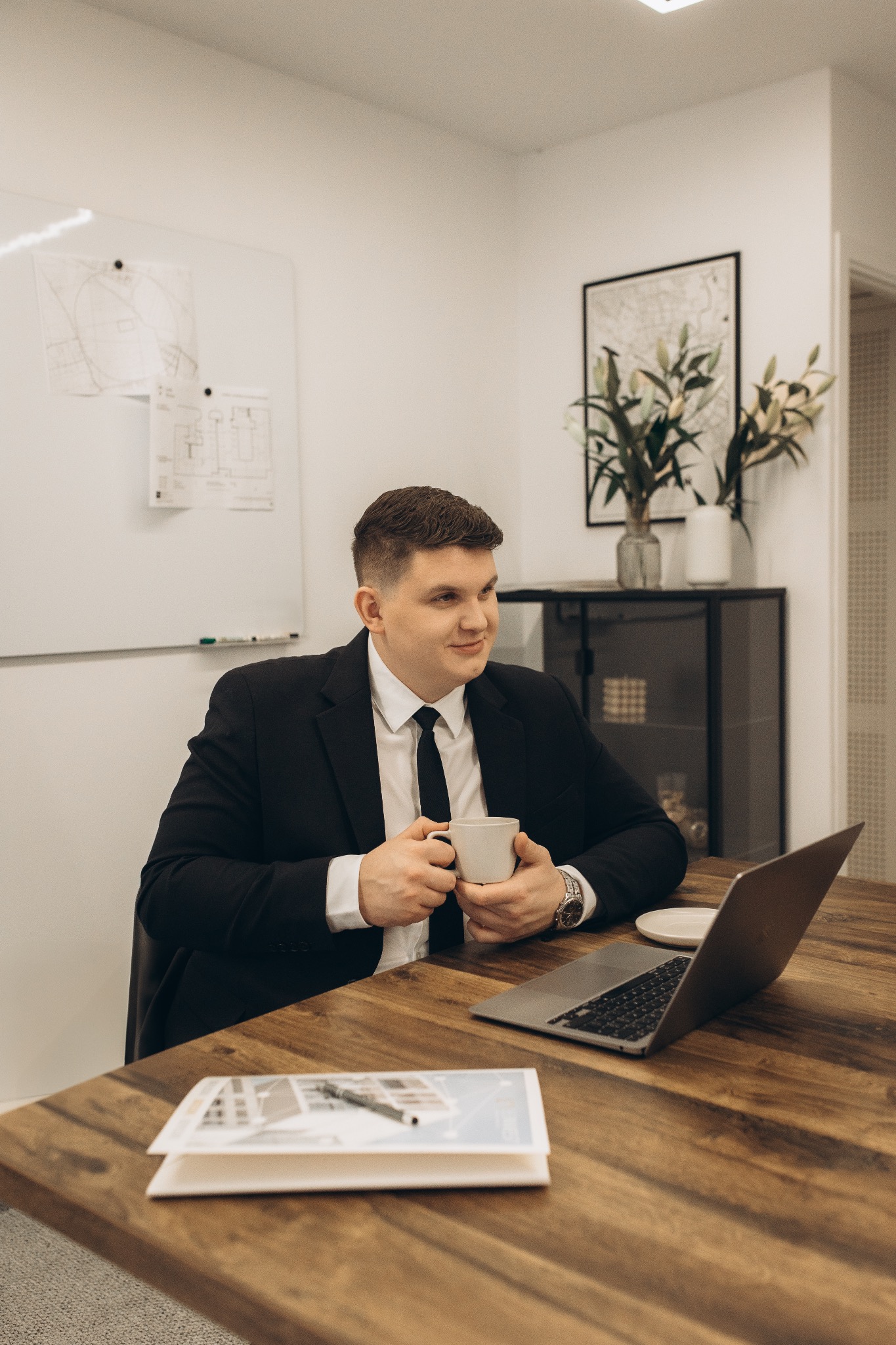 Agent nieruchomości przy biurku z laptopem i planami, pije kawę. Na ścianie mapa i rysunki architektoniczne. Uśmiechnięty, w garniturze. Profesjonalne biuro.