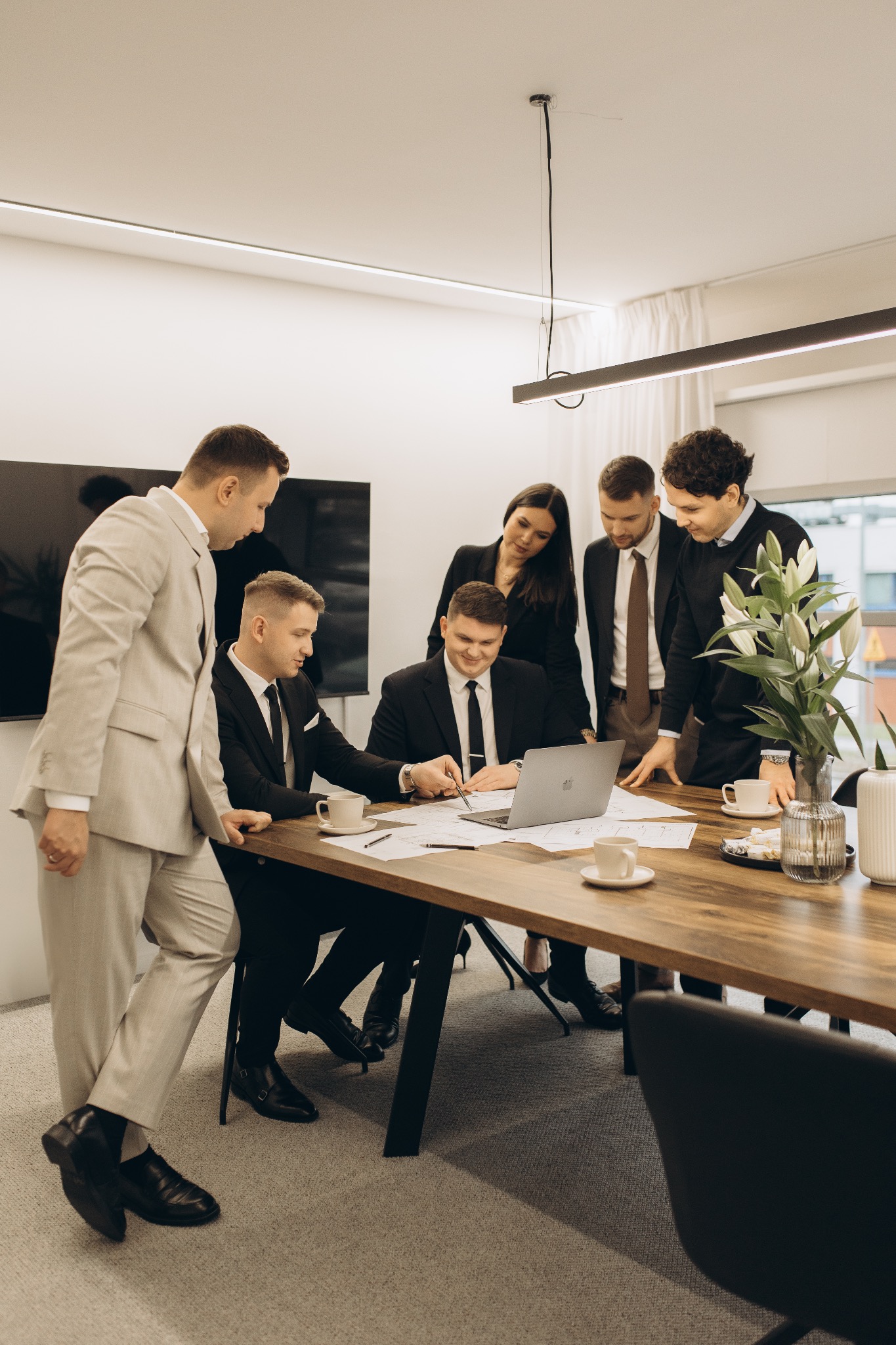 Zespół w biurze analizuje dokumenty przy drewnianym stole z laptopem i kawą. Jasne wnętrze, nowoczesne oświetlenie. Jeden z mężczyzn w beżowym garniturze.