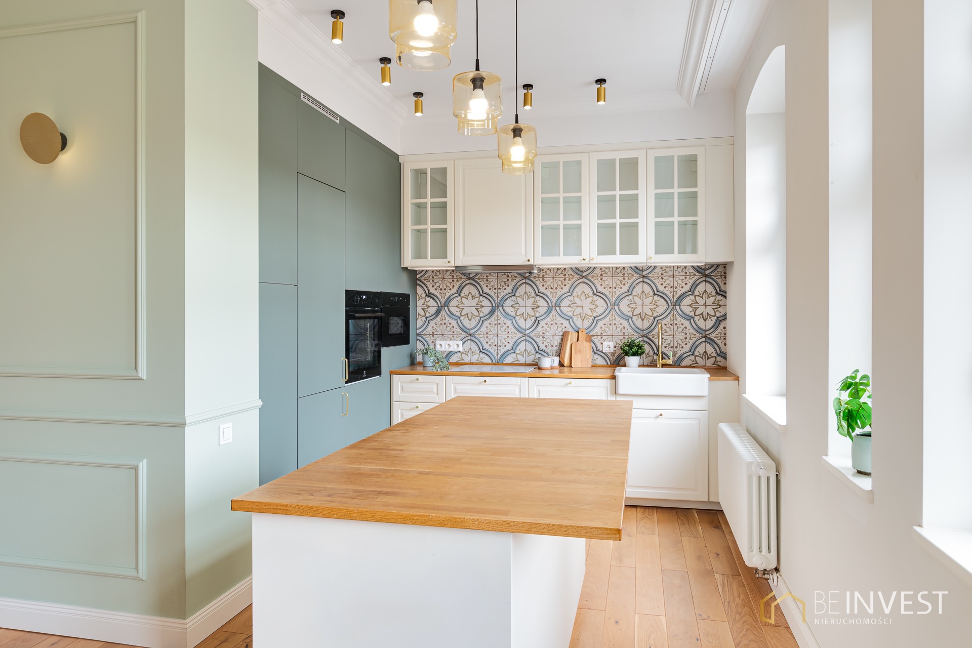 Elegancka kuchnia w stylu retro z wyspą, mozaiką nad blatem i złotymi detalami oświetlenia. Pastelowe kolory ścian i zabudowy, drewniana podłoga.