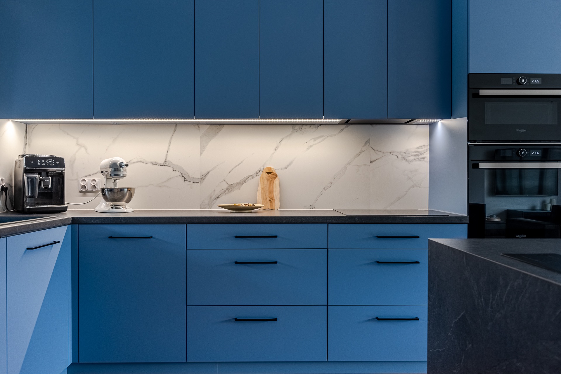 Niebieska kuchnia z marmurowym backsplash, oświetleniem LED i sprzętami AGD: ekspres do kawy, mikser planetarny i piekarnik w zabudowie. Nowoczesny design.