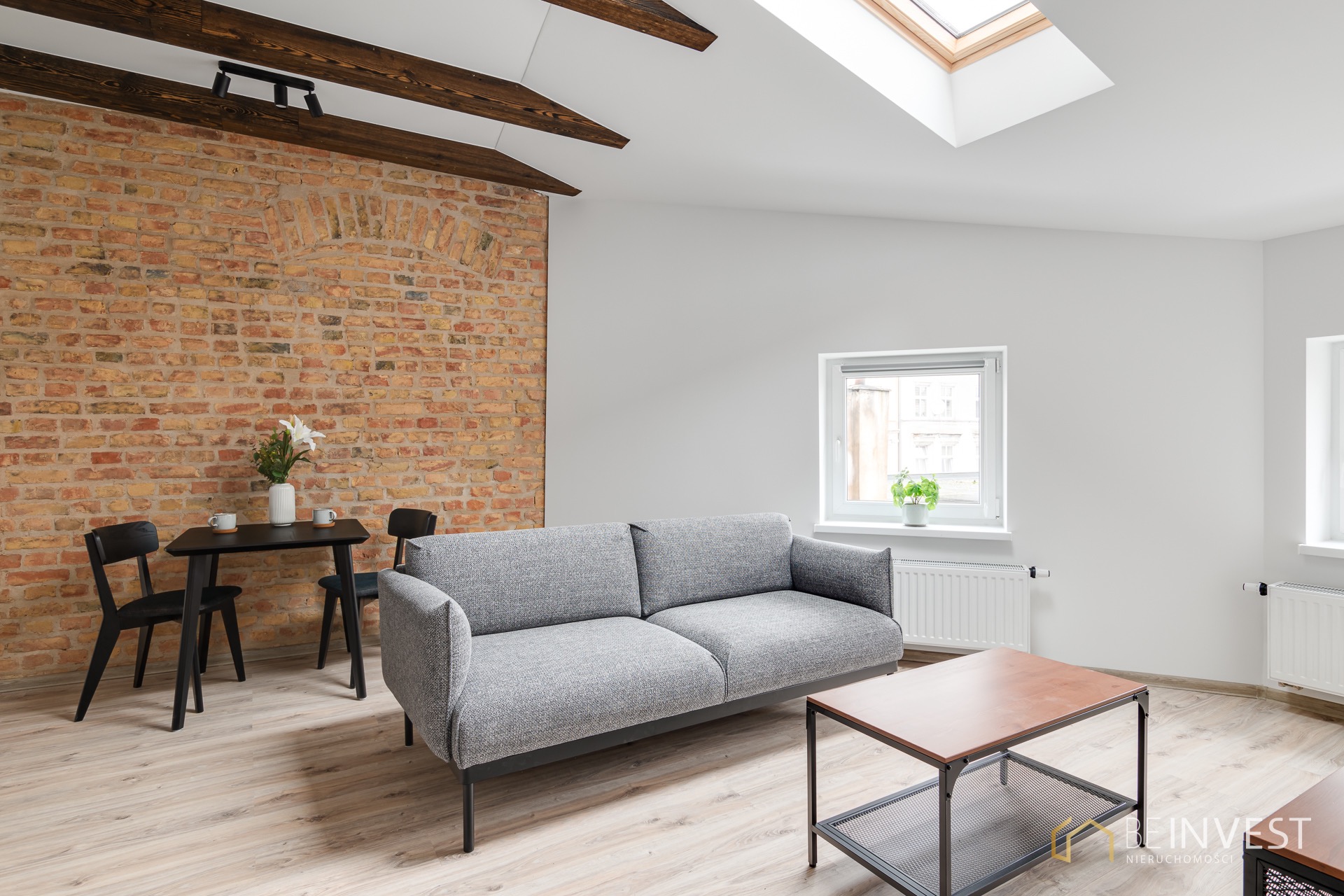 Salon z szarą sofą, ceglaną ścianą i drewnianymi belkami. Widoczne okno dachowe, stół z krzesłami i stolik kawowy. Jasne wnętrze, minimalistyczny design.