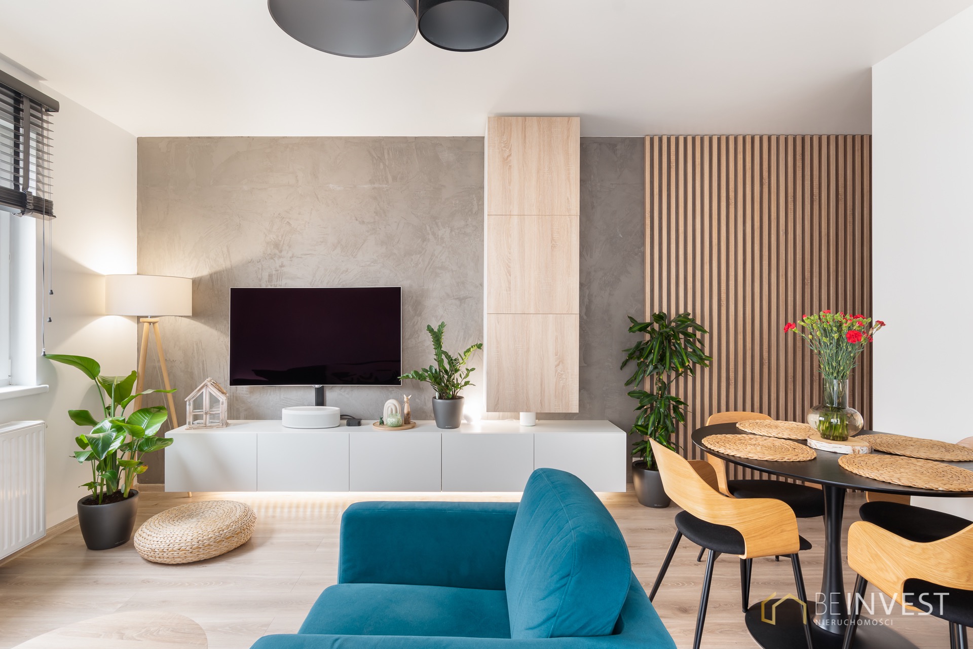 Nowoczesny salon z turkusową sofą, minimalistyczną szafką RTV podświetloną LED, drewnianymi lamelami na ścianie i okrągłym stołem z kwiatami.