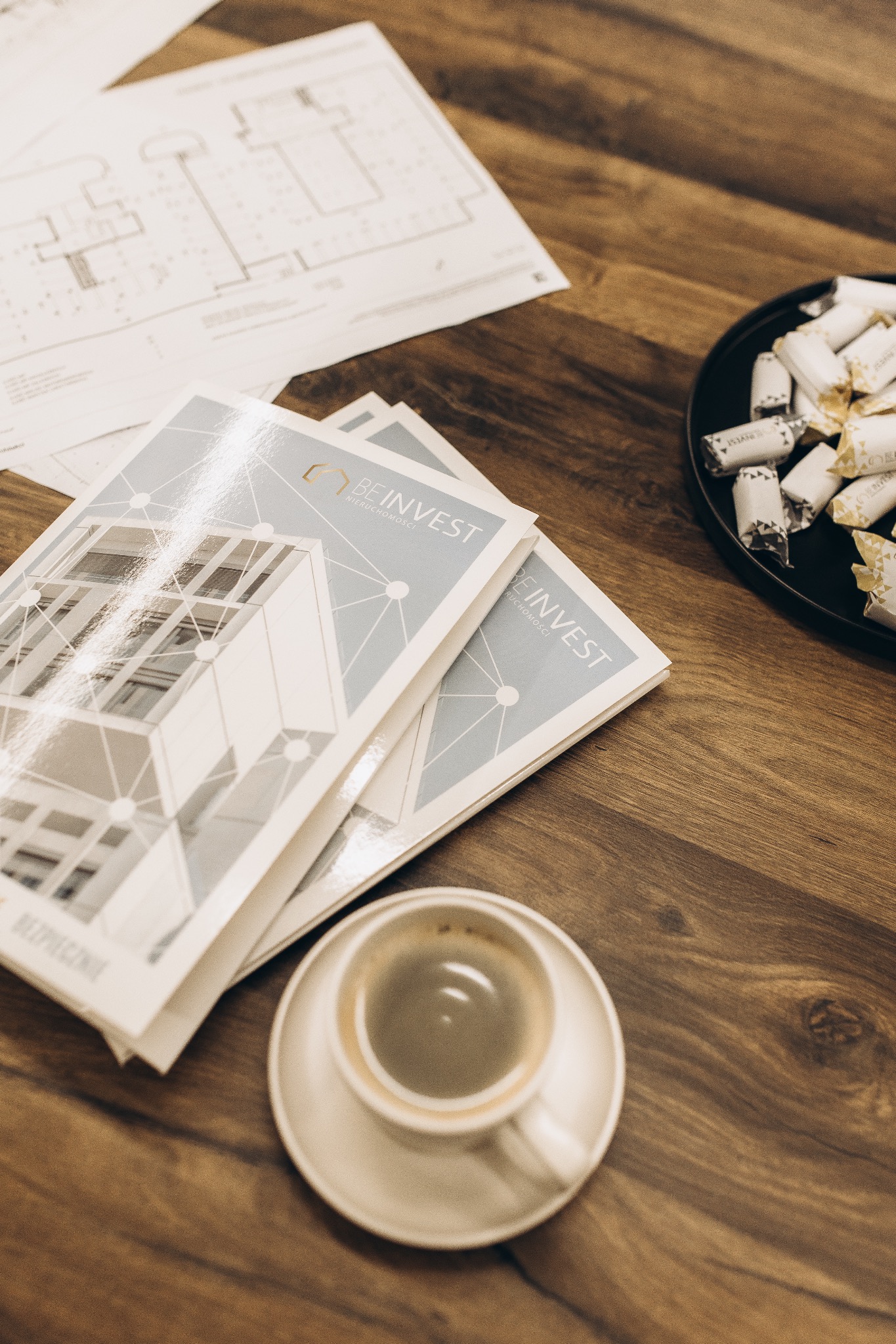 Broszury 'Be Invest Nieruchomości' na drewnianym stole z kawą i słodyczami. Widoczny plan budynku. Ciepłe oświetlenie.