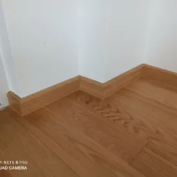 Jasne drewniane panele podłogowe ułożone w rogu pomieszczenia, z dopasowanymi listwami przypodłogowymi w kolorze drewna, przylegającymi do białej ściany.