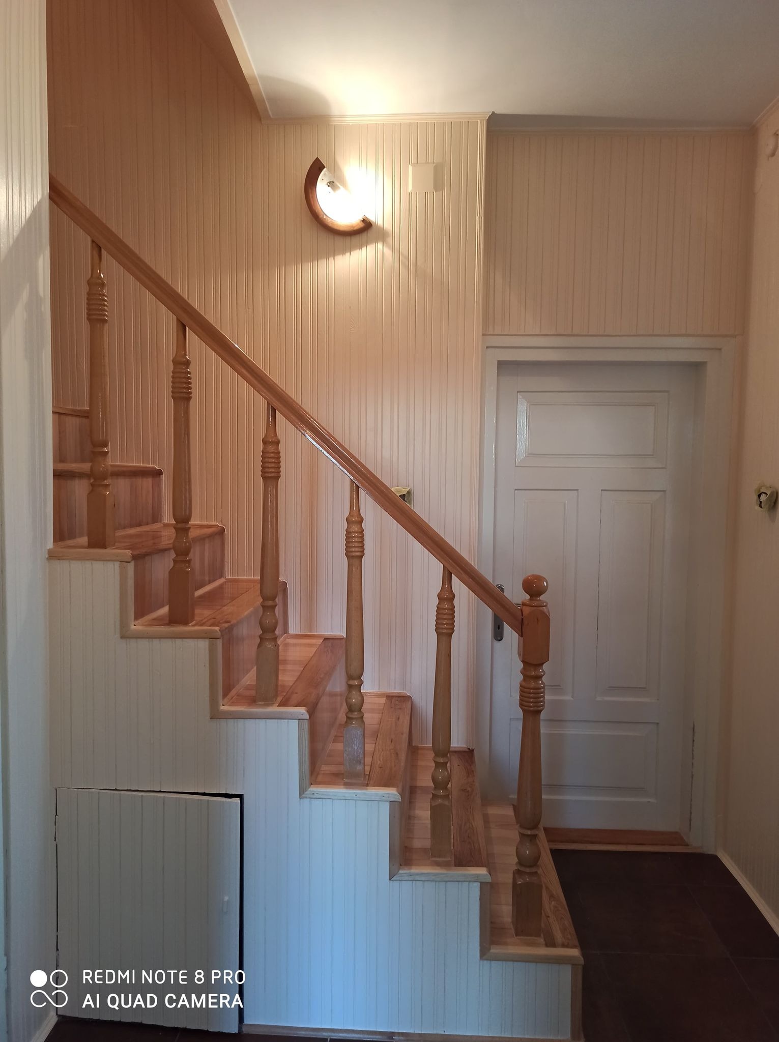 Drewniane schody z balustradą w jasnym wnętrzu, z widoczną lampą ścienną i drzwiami w tle.