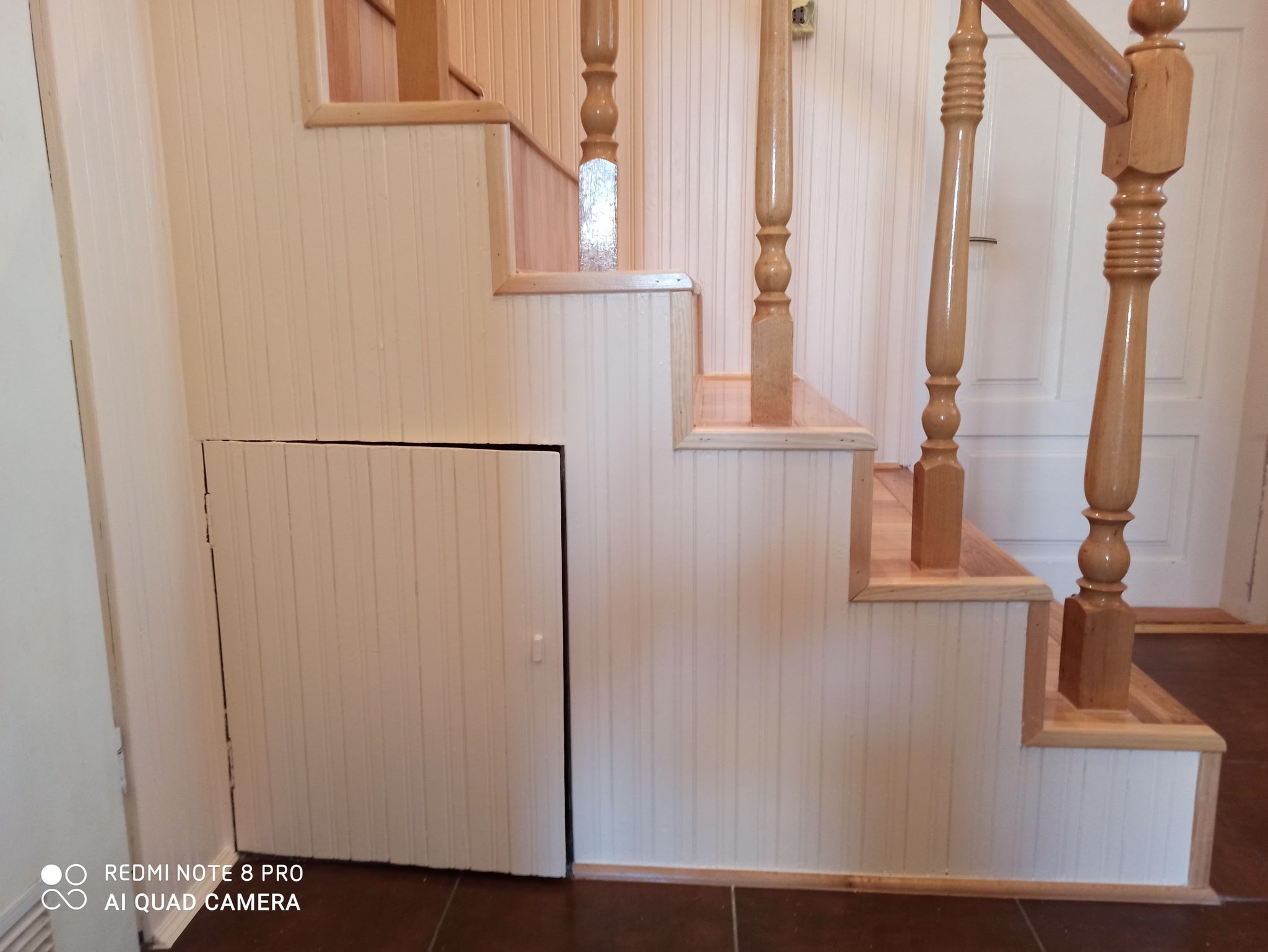 Jasne, drewniane schody z ozdobnymi balustradami i ukrytą szafką pod spodem, wykończone panelami ściennymi z pionowymi żłobieniami.