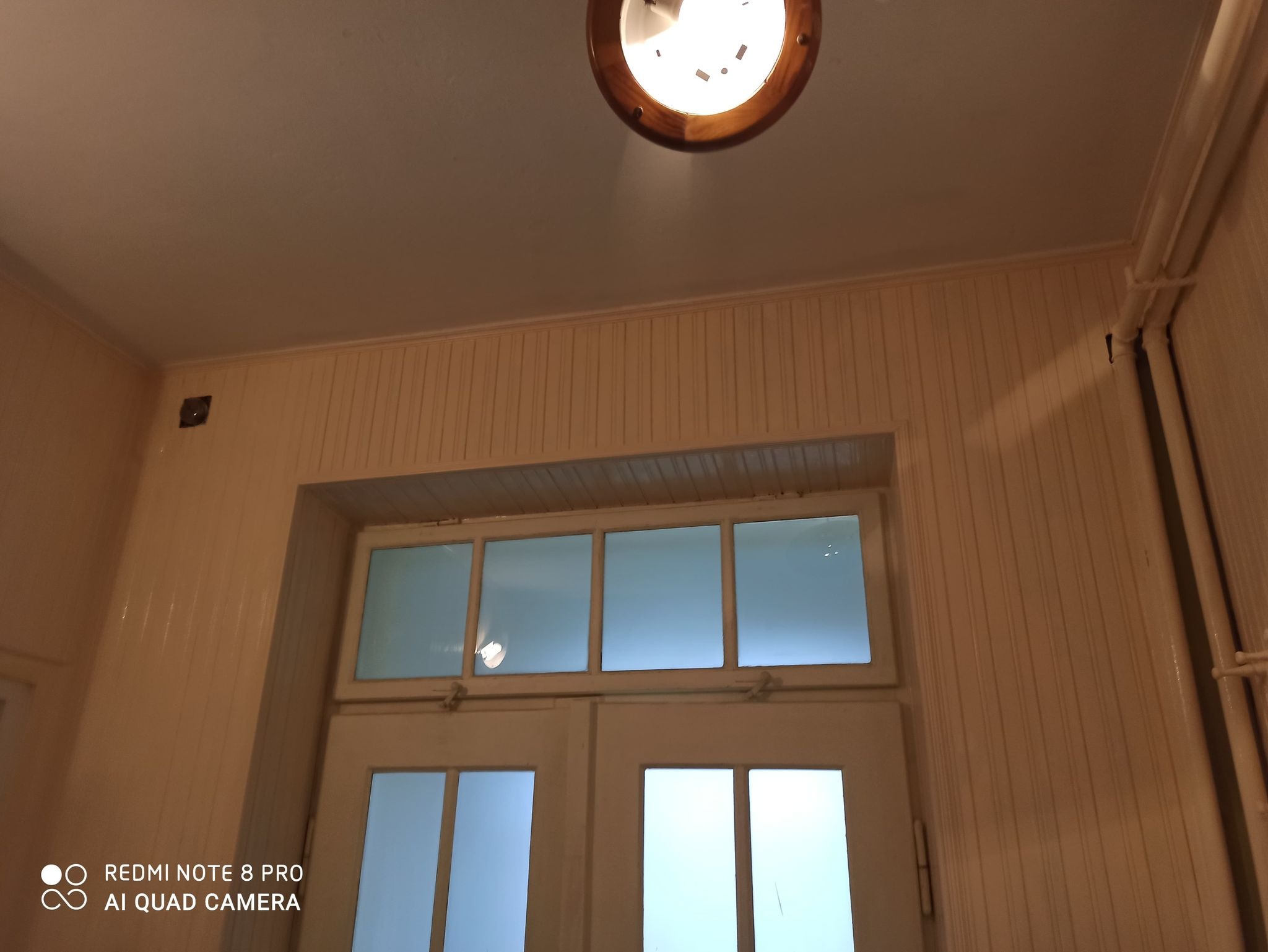 Wnętrze przedpokoju z boazerią w odcieniach beżu, przeszklone drzwi i okno nad nimi, oświetlenie sufitowe w stylu retro.