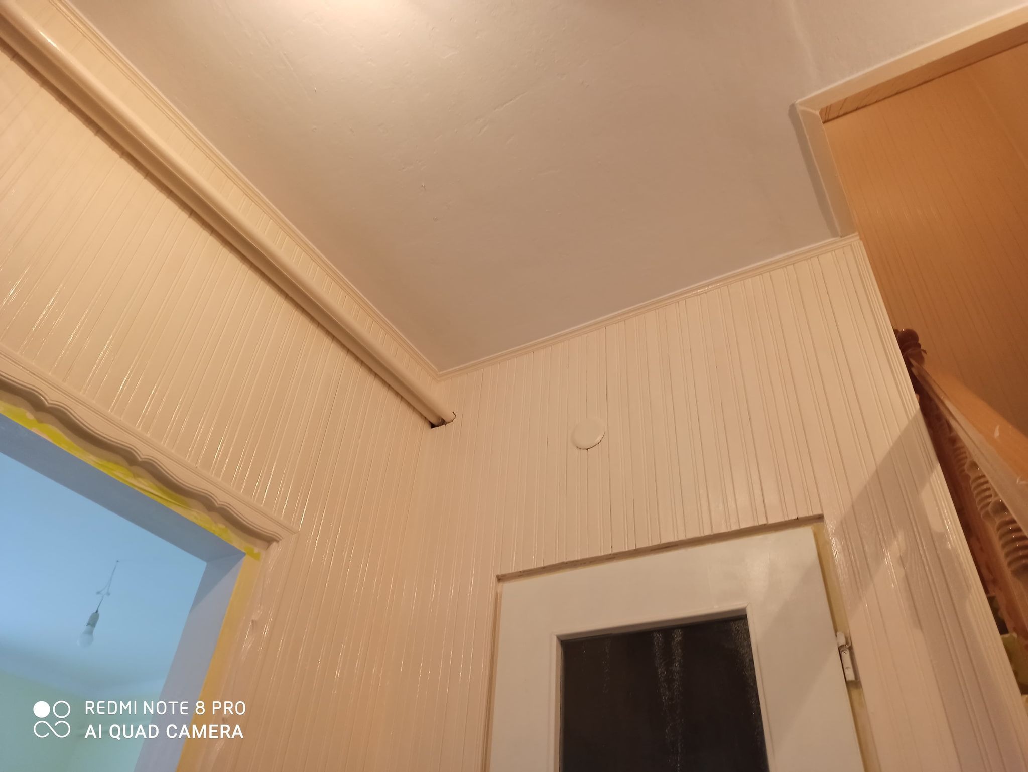 Wykończona ściana z panelami boazeryjnymi w odcieniach beżu, widoczne listwy przypodłogowe, framuga drzwi i fragment schodów z drewnianą balustradą, ujęcie z dołu.