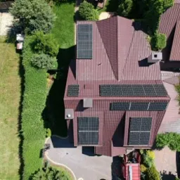 Widok z góry na dom z brązowym dachem, na którym zamontowane są panele słoneczne. W tle widoczna zielona trawa i drzewa. Częściowo widoczny samochód osobowy.