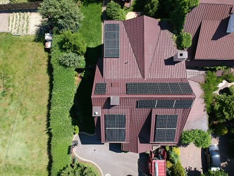 Widok z góry na dom z brązowym dachem, na którym zamontowane są panele słoneczne. W tle widoczna zielona trawa i drzewa. Częściowo widoczny samochód osobowy.