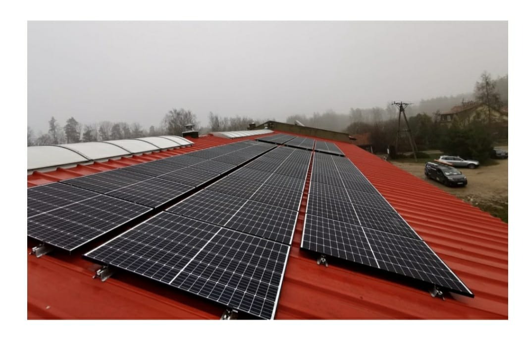 Instalacja paneli słonecznych na czerwonym dachu budynku w pochmurny dzień, widoczne samochody w tle.