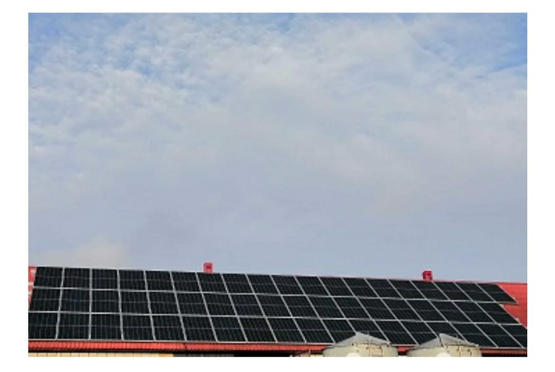 Czarny układ paneli słonecznych na czerwonym dachu budynku, widok z dołu, z fragmentem nieba z chmurami w tle.