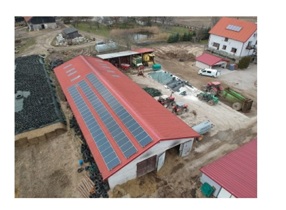 Widok z góry na duży budynek gospodarczy z czerwonym dachem, na którym zamontowane są panele słoneczne w trzech rzędach. Obok sterta opon i maszyny rolnicze.