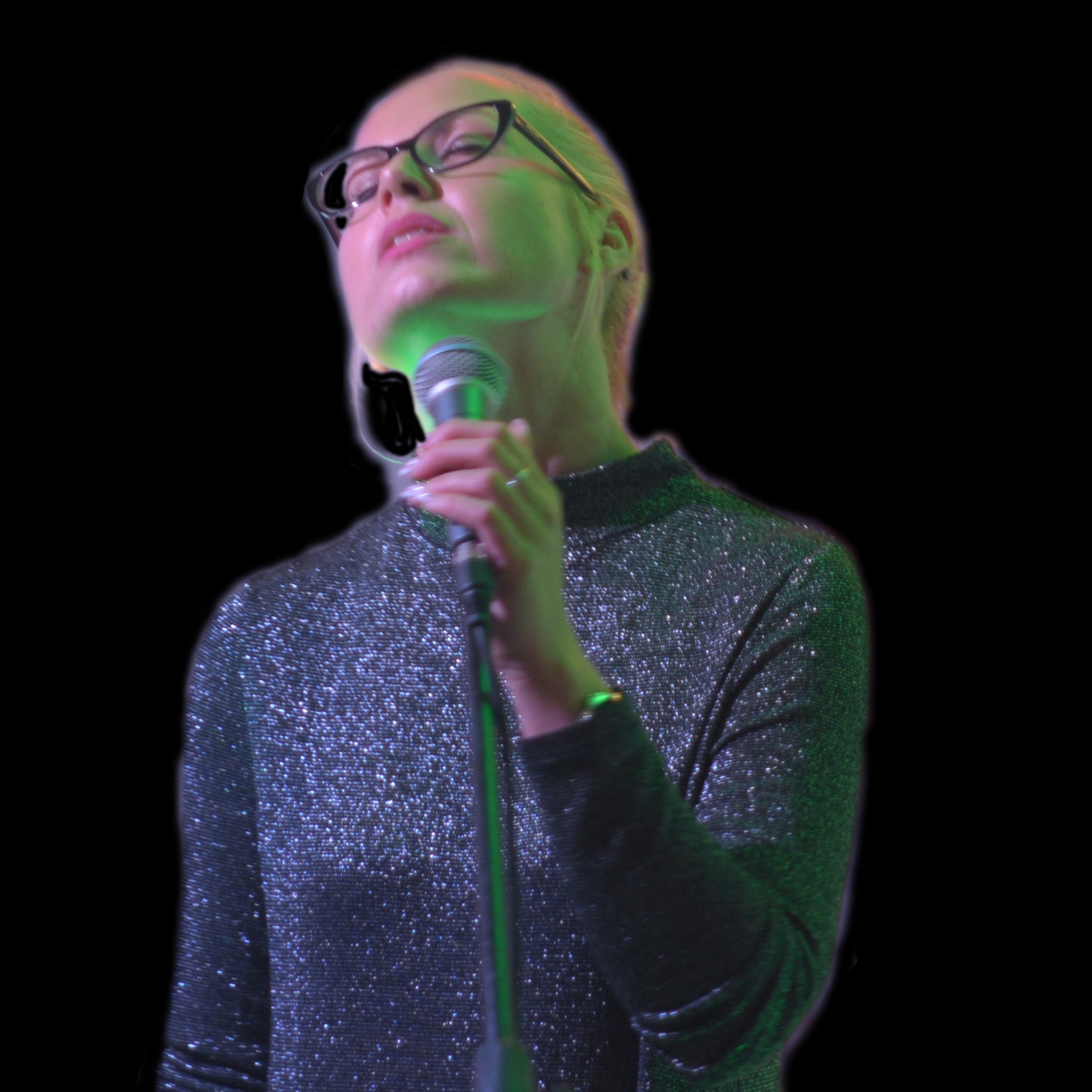 Wokalistka z Bydgoszczy w błyszczącej sukience, śpiewająca do mikrofonu oświetlonego zielonym światłem, z czarnymi okularami na nosie.