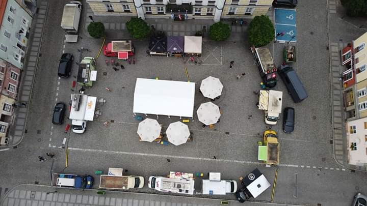 Widok z lotu ptaka na rynek miejski z rozstawionymi stoiskami gastronomicznymi, namiotem i parasolami, otoczony budynkami i ulicami.