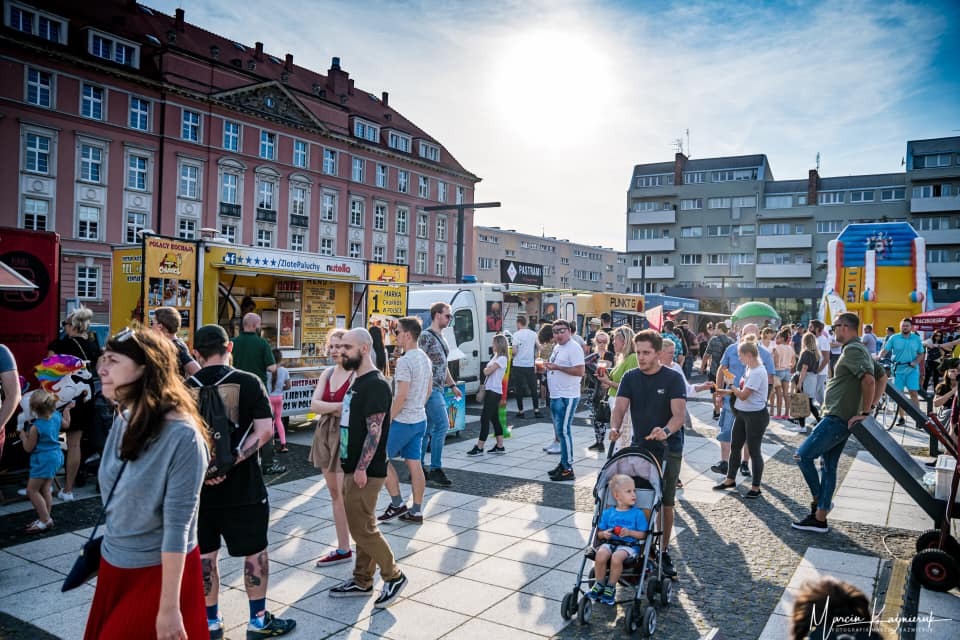 Tłum ludzi na placu miejskim podczas festynu z food truckami i dmuchańcem w tle.