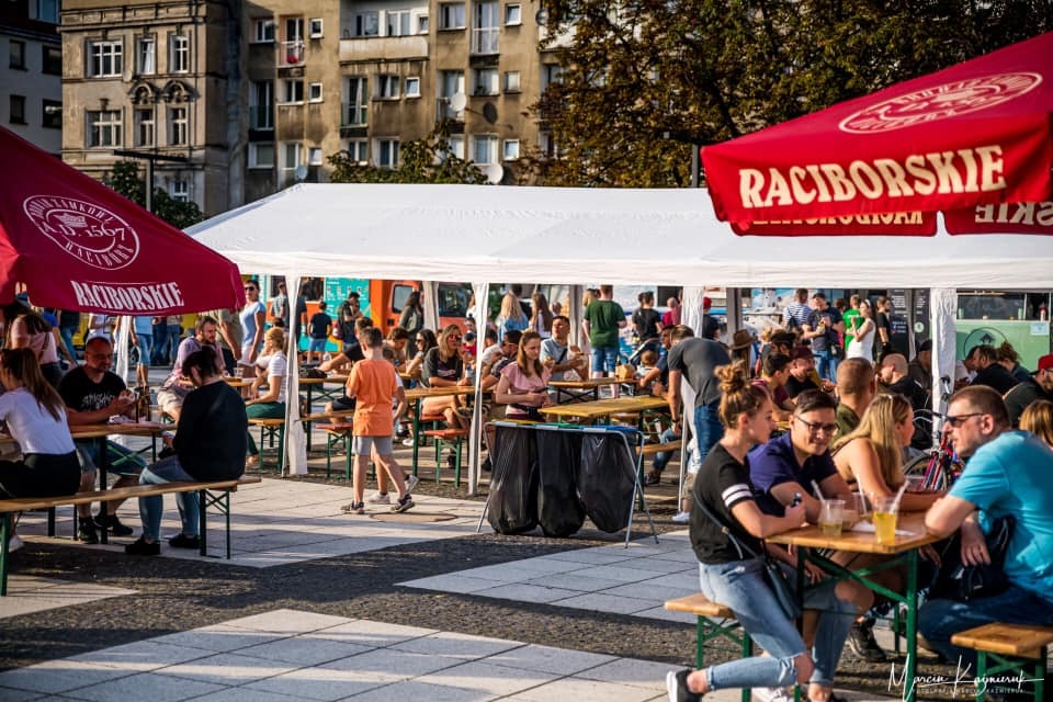 Plenerowa impreza z białym namiotem, drewnianymi stołami i ławkami, parasolami z logo 'Raciborskie', licznymi gośćmi w tle miejska zabudowa.
