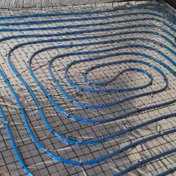 Niebieskie rury grzewcze ułożone spiralnie na srebrnej folii z czarną siatką, mocowane plastikowymi klipsami, widok z góry na surową podłogę przed wylewką.