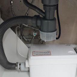 Dariusz Wiśniewski - Szara karbowana rura odpływowa podłączona do białej pompy sanitarnej w szafce pod zlewem, widoczne również rury doprowadzające wodę z zaworami.