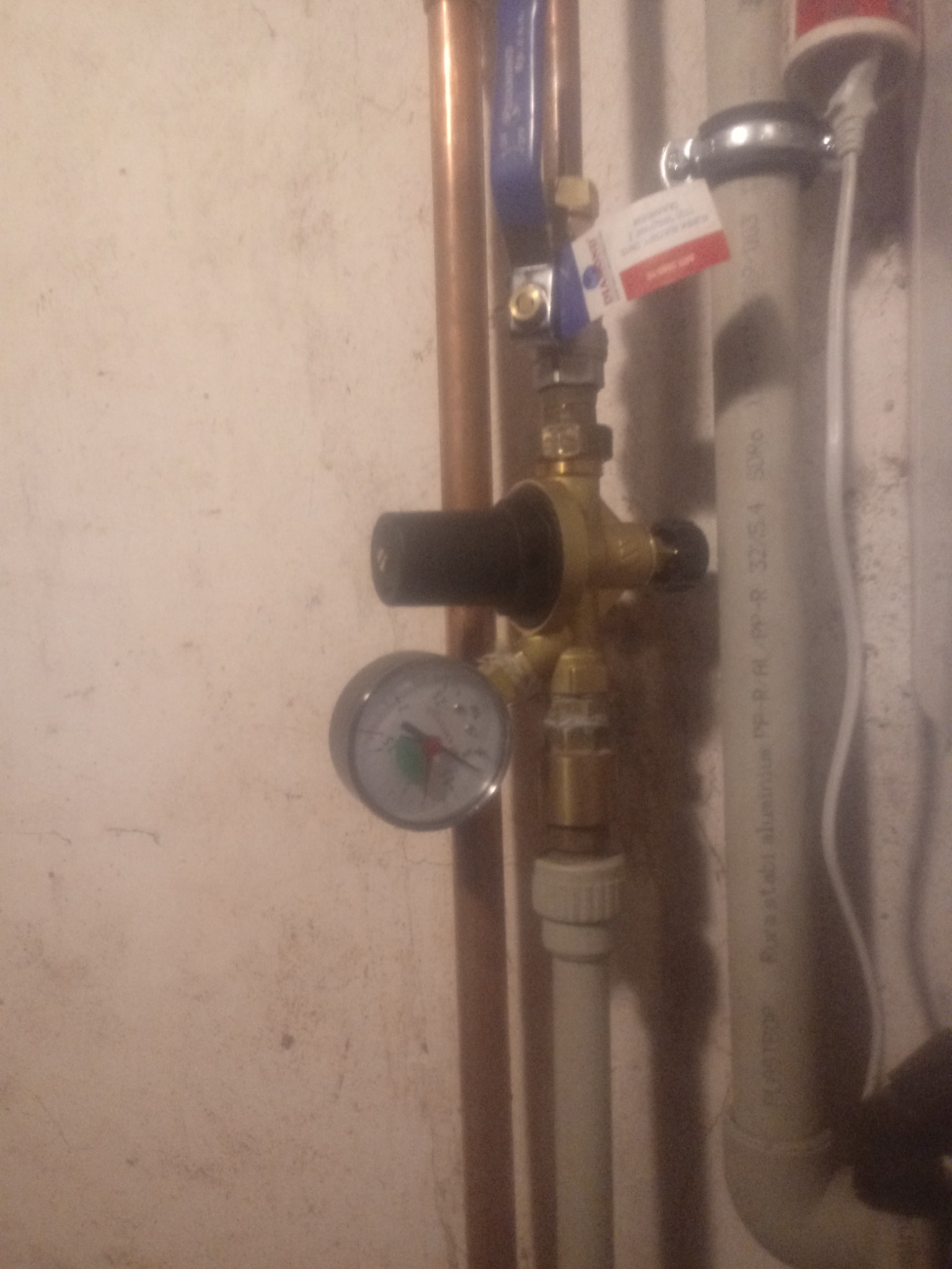 Manometr wskazujący ciśnienie w instalacji, zawór kulowy, rury miedziane i plastikowe, widoczne przyłącza i adaptery.