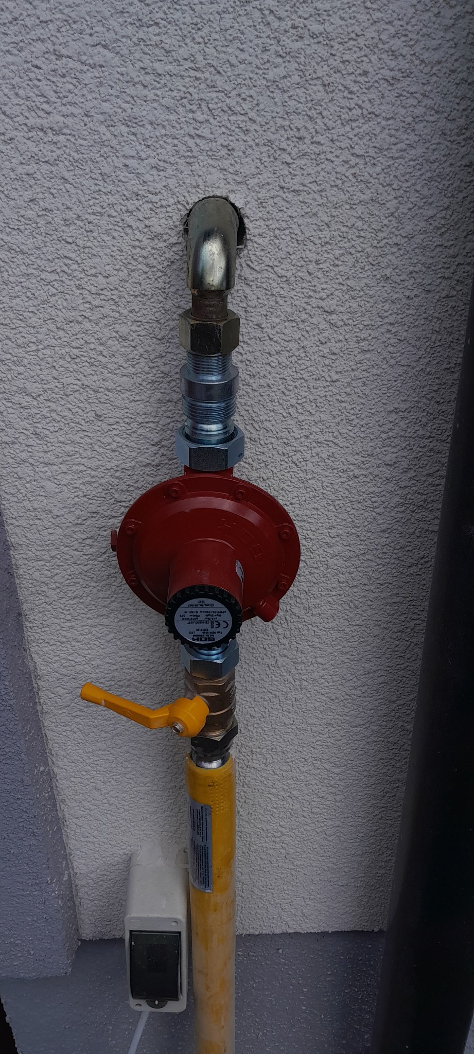 Czerwony reduktor gazu z żółtym zaworem na żółtej rurze, zamontowany na szarej ścianie w Grodzisku Mazowieckim.