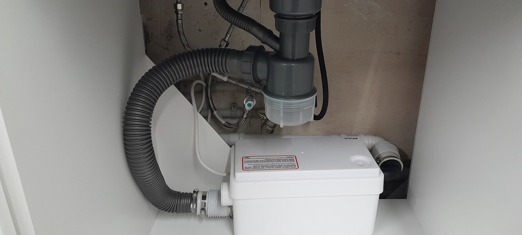 Szara karbowana rura odpływowa podłączona do białej pompy sanitarnej w szafce pod zlewem, widoczne również rury doprowadzające wodę z zaworami.