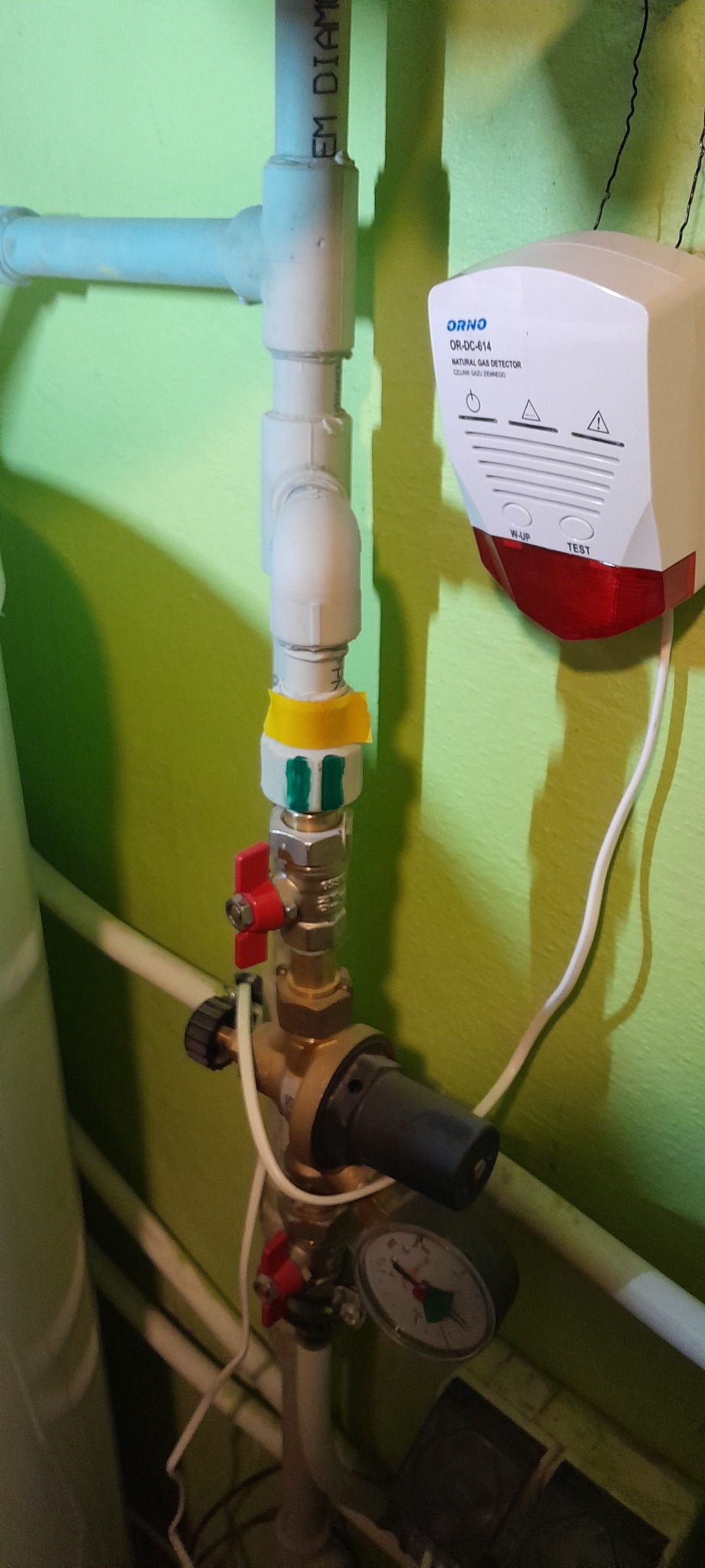 Fragment instalacji gazowej z detektorem gazu Orno OR-DC-614 na tle zielonej ściany, widoczne rury, zawory, manometr i elementy z żółtą taśmą izolacyjną.