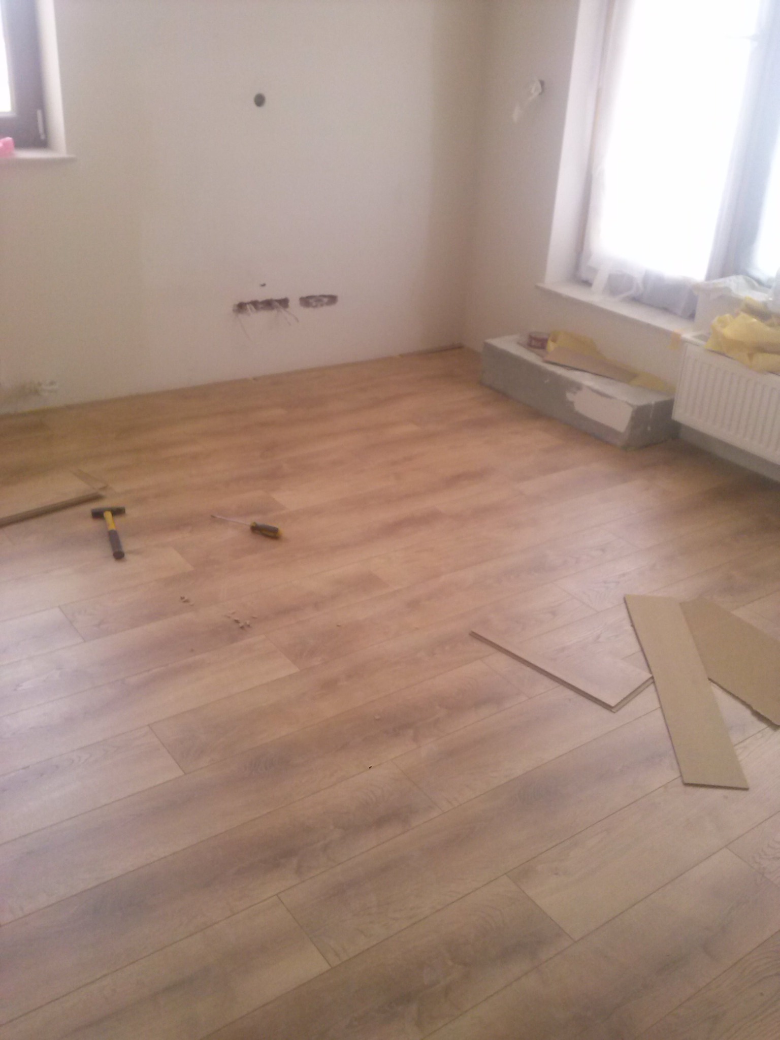 Układanie paneli podłogowych w pokoju z widocznymi narzędziami: młotek i śrubokręt, oraz paczką paneli przy grzejniku.