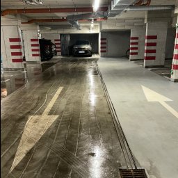 BŁYSK SERVICE USŁUGI PORZĄDKOWE - Mokry parking podziemny po umyciu posadzki. Widoczne ślady opon i biała strzałka kierunkowa. Część parkingu sucha, część mokra. Słupy z czerwonymi pasami.