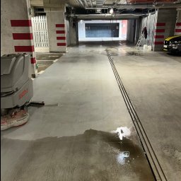 BŁYSK SERVICE USŁUGI PORZĄDKOWE - Maszyna czyszcząca Comac na mokrej posadzce parkingu podziemnego. Widoczne ślady po czyszczeniu, kolumny z czerwonymi pasami, rury pod sufitem i wyjście ewakuacyjne.