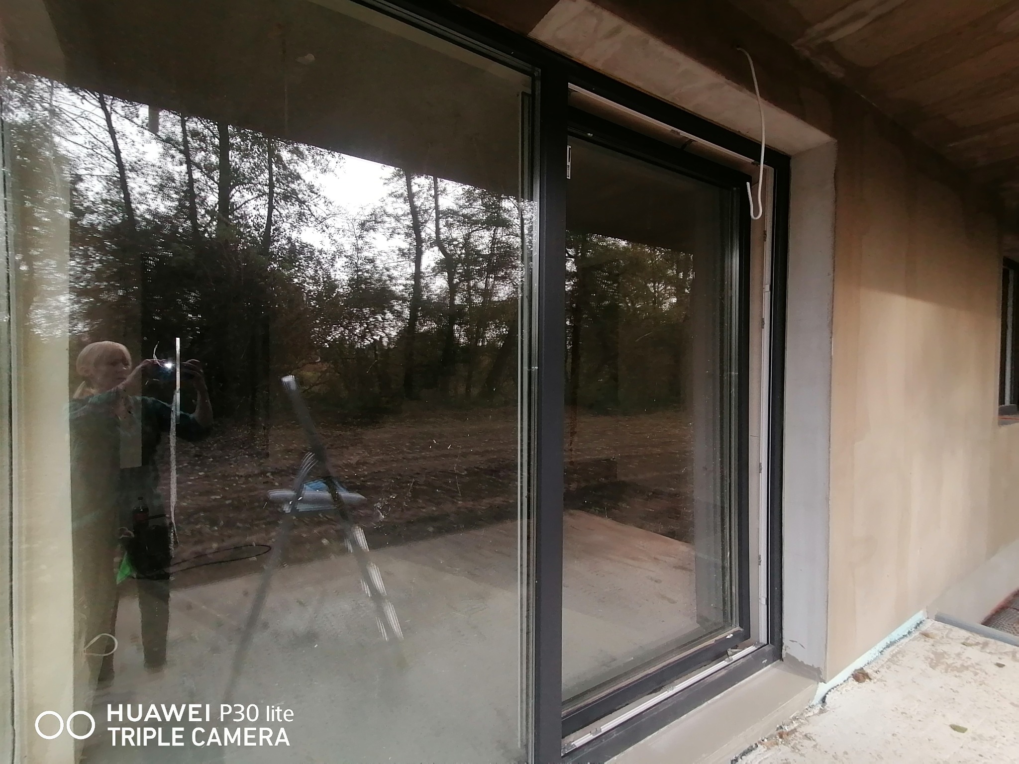 Duże przesuwne okno z widocznym odbiciem otoczenia, drabina w tle, surowe wykończenie ściany zewnętrznej, ślady po budowie.