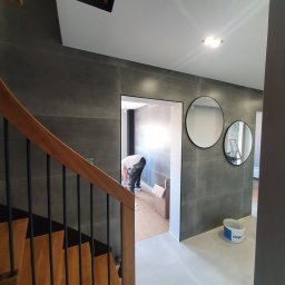 Denis Popiński - Korytarz w trakcie prac wykończeniowych: szare płytki wielkoformatowe na ścianach, dwa okrągłe lustra, drewniane schody z czarnymi balustradami, pracownik w tle przygotowuje podłoże, widoczne...