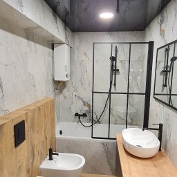 Denis Popiński - Nowoczesna łazienka z marmurowymi płytkami, czarną armaturą i drewnianymi akcentami. Uwagę przykuwa podwieszana toaleta i bidet oraz owalna umywalka na drewnianym blacie.