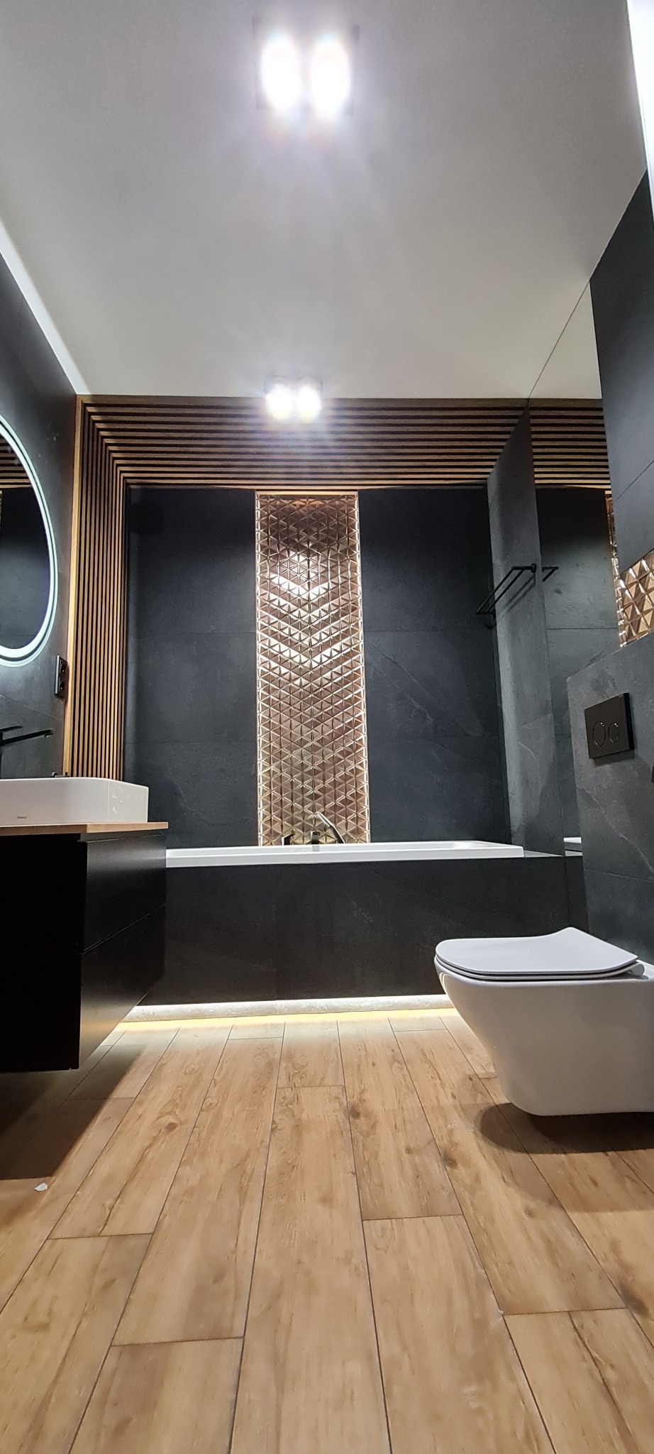Elegancka łazienka z drewnianymi akcentami i czarnymi płytkami, subtelne oświetlenie LED pod wanną i oryginalna mozaika. Nowoczesny design.
