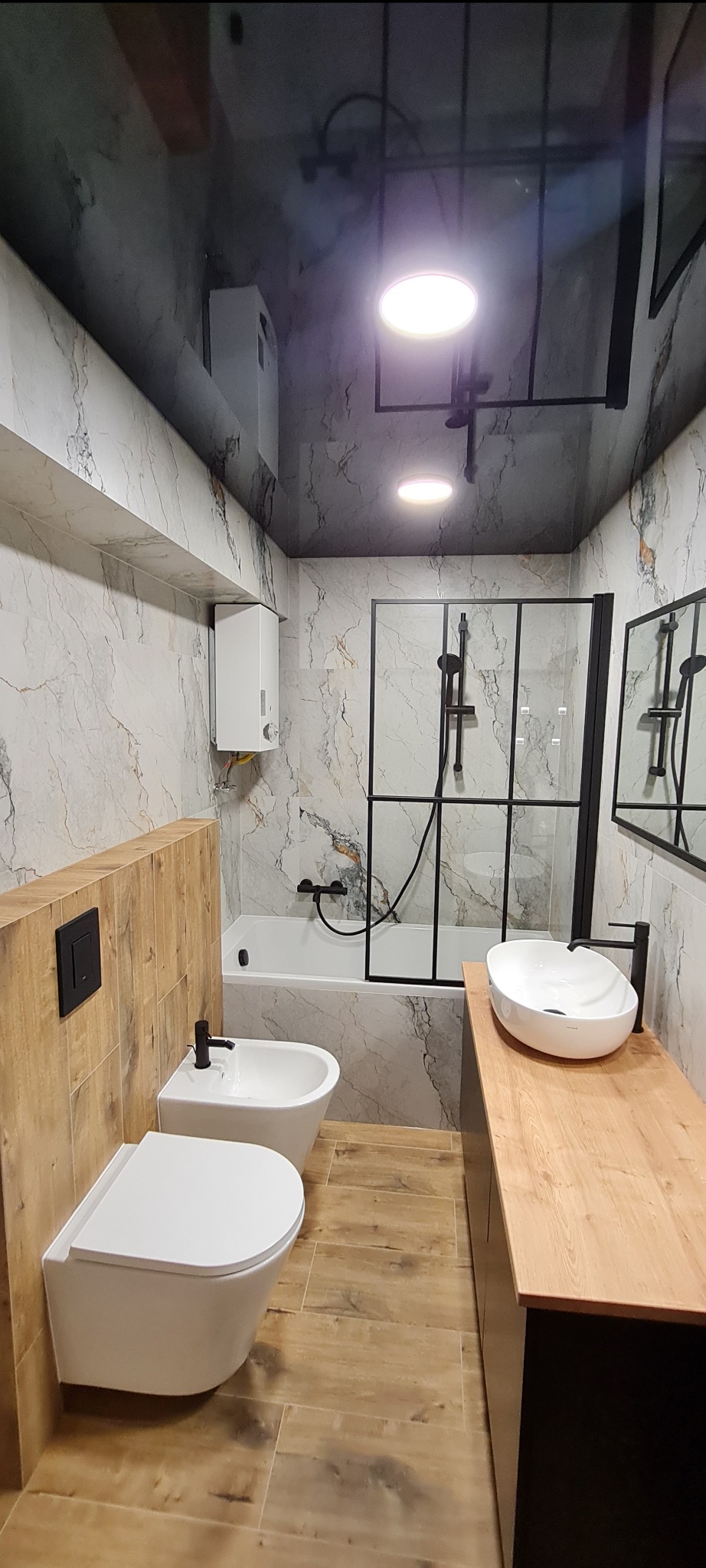 Nowoczesna łazienka z marmurowymi płytkami, czarną armaturą i drewnianymi akcentami. Uwagę przykuwa podwieszana toaleta i bidet oraz owalna umywalka na drewnianym blacie.